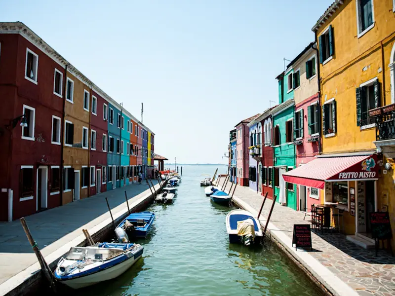 Case colorate e barche lungo un canale di Burano aperto verso la laguna, sotto un cielo azzurro