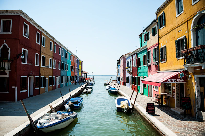 Case colorate e barche lungo un canale di Burano aperto verso la laguna, sotto un cielo azzurro